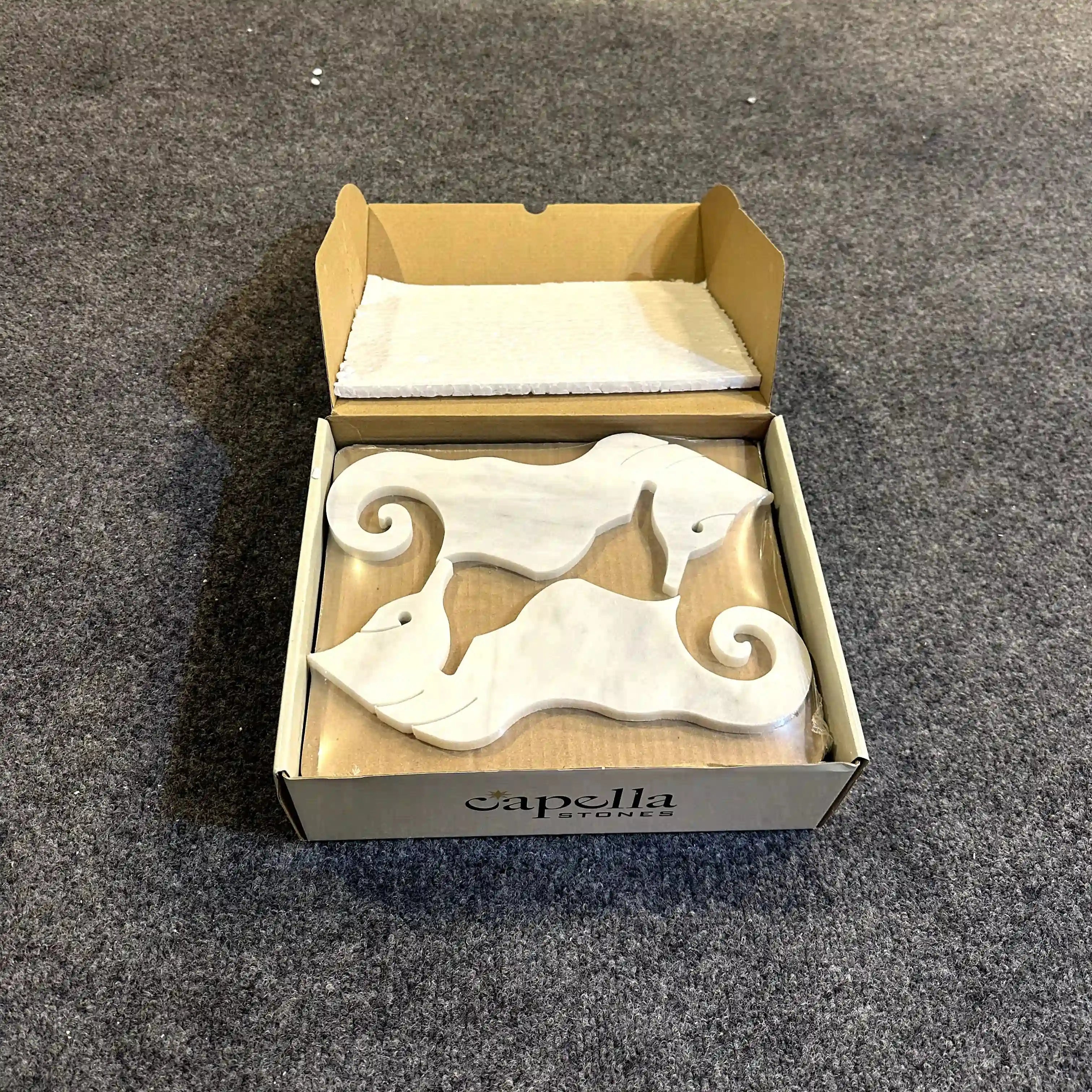 Two white ceramic dog figurines in a 'capella' branded box on a gray carpet.