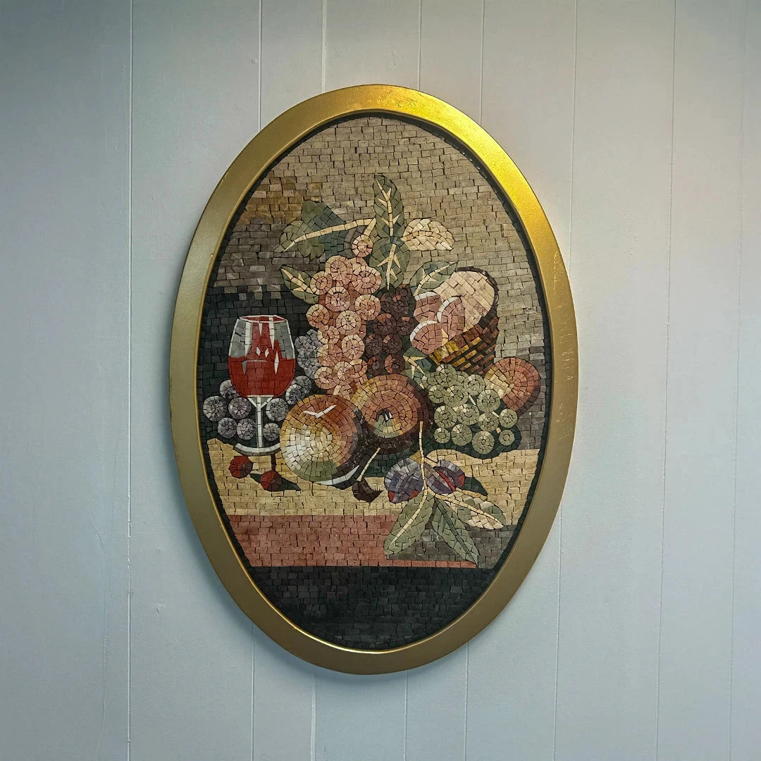 Mosaic artwork of a still life scene with fruits and a glass of wine, framed in gold, on a light blue wall.