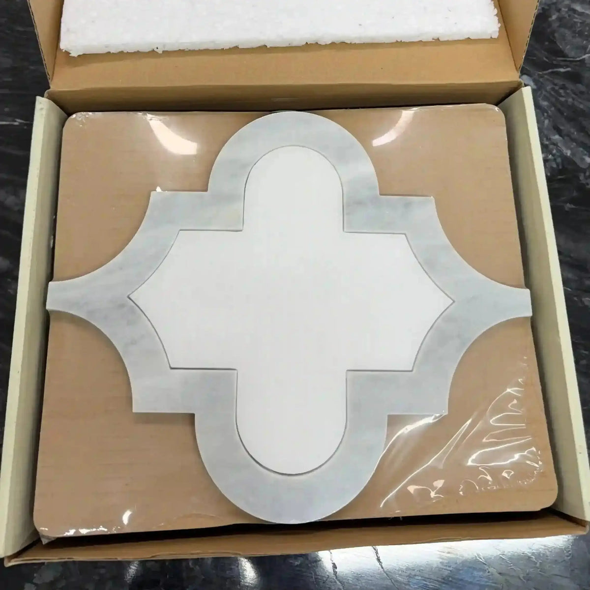 Ares natural marble mosaic tile packaged securely inside a cardboard shipping box, featuring an ornate, cross-like quatrefoil architectural silhouette with a smooth white marble center framed by a contrasting gray marble border.