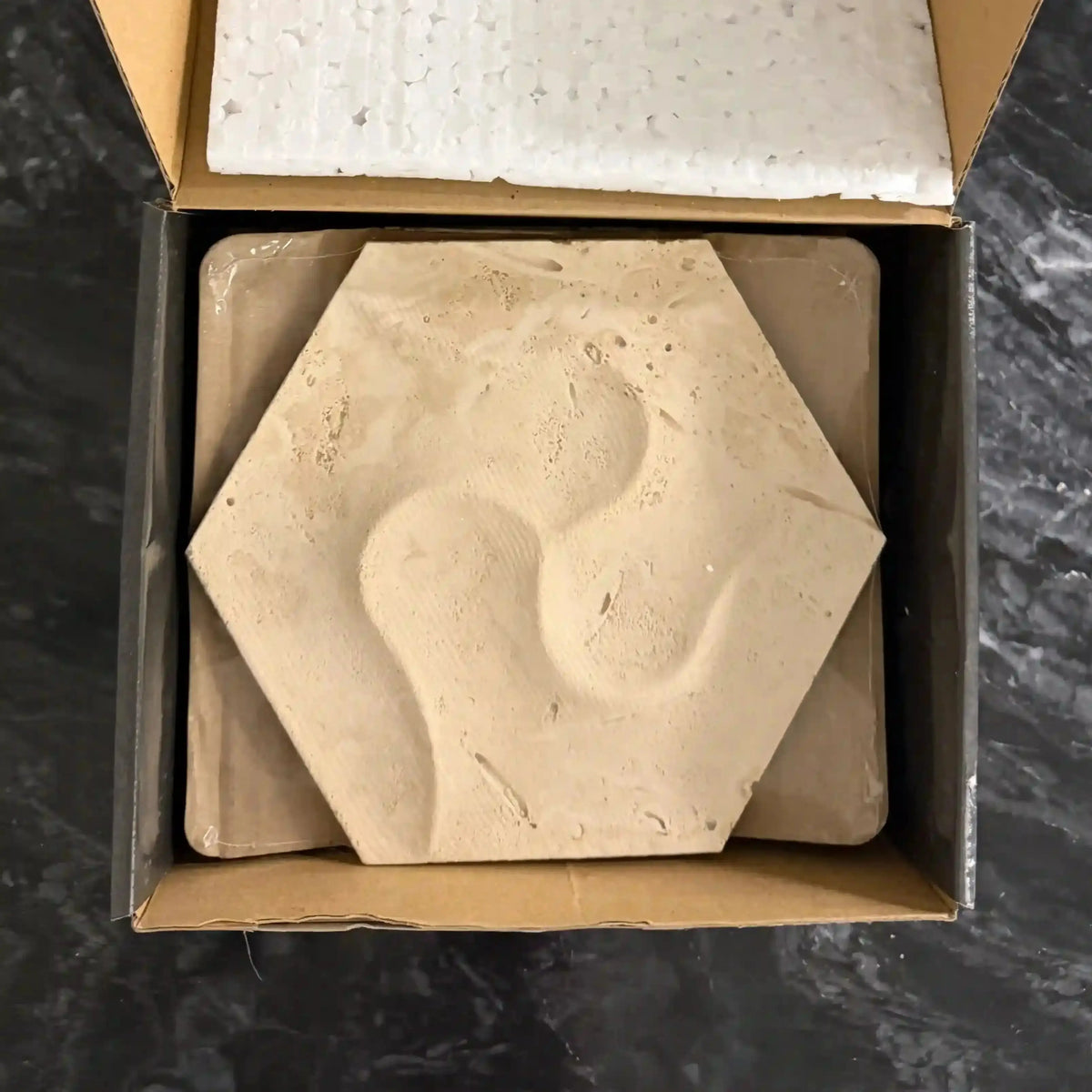 Calypso natural stone 3D wall tile packaged neatly inside a cardboard box with protective foam lining, showcasing a sculptural hexagonal design with fluid, organic concave indentations in a warm beige travertine finish.
