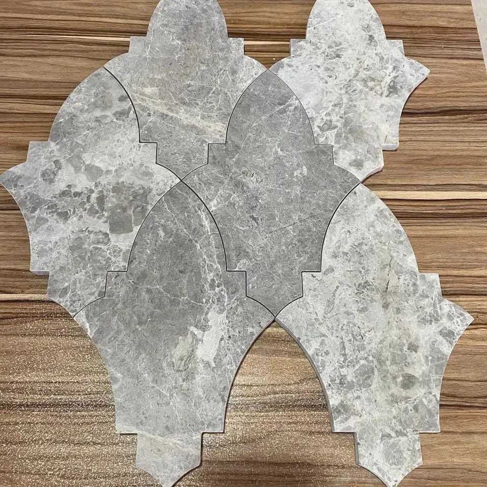 A close-up arrangement of Khepri marble mosaic tiles displayed on a warm wooden surface, showcasing the detailed grey natural stone grain and unique interlocking lantern-like shape.