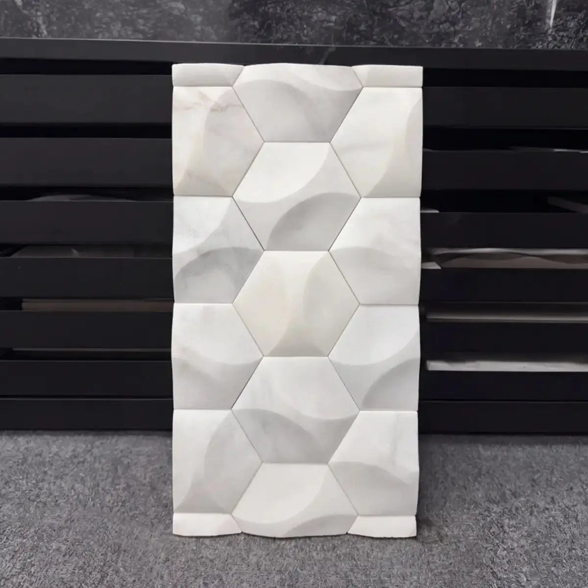 Orbis natural stone 3D wall tile sample displayed vertically against a dark background, showcasing its repeating geometric hexagonal pattern with concave carved facets and a smooth polished white marble surface.