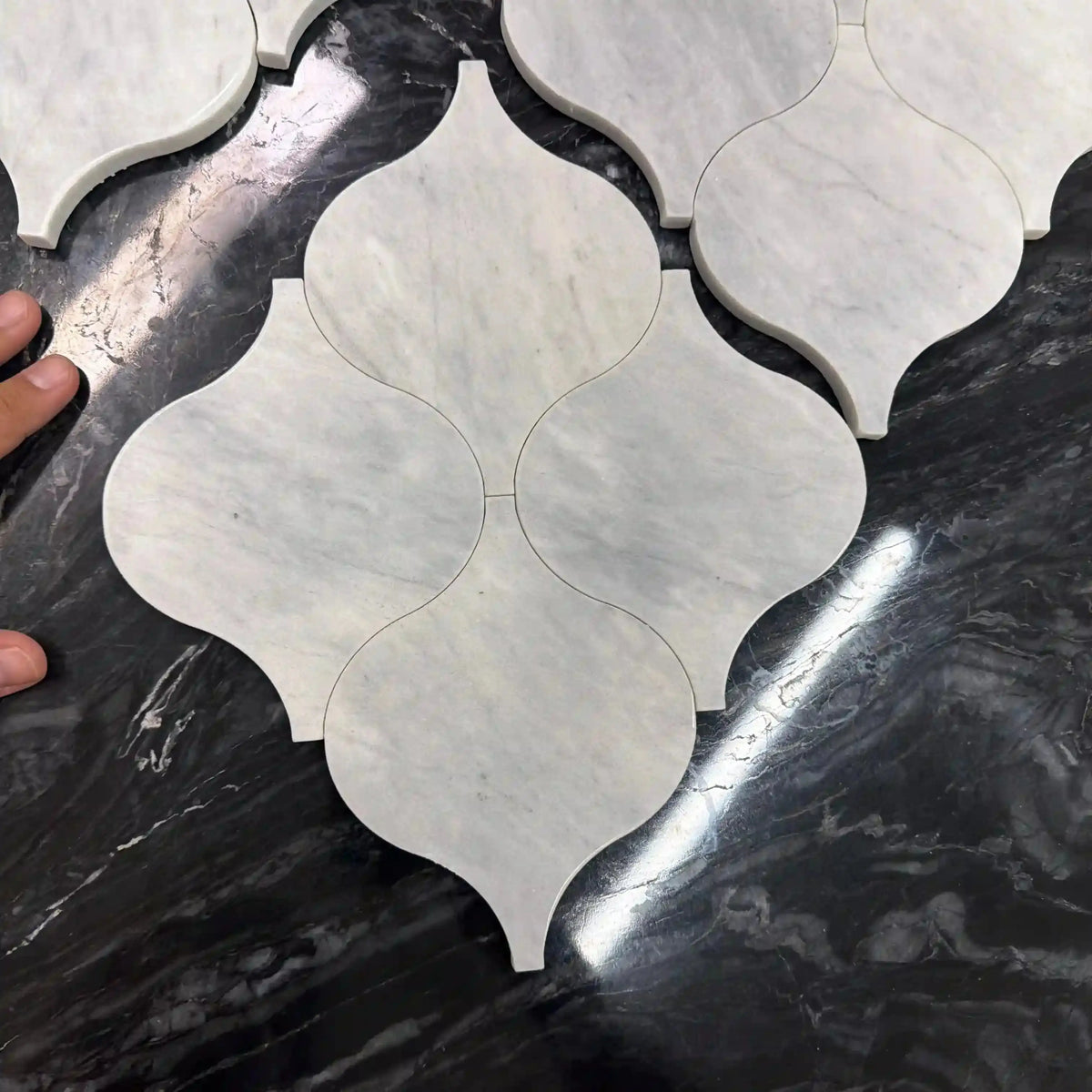 Edrice natural stone mosaic tile close-up view highlighting the interlocking curved, teardrop-shaped white marble pieces with subtle gray veining on a dark marble surface, with a hand visible on the left edge.
