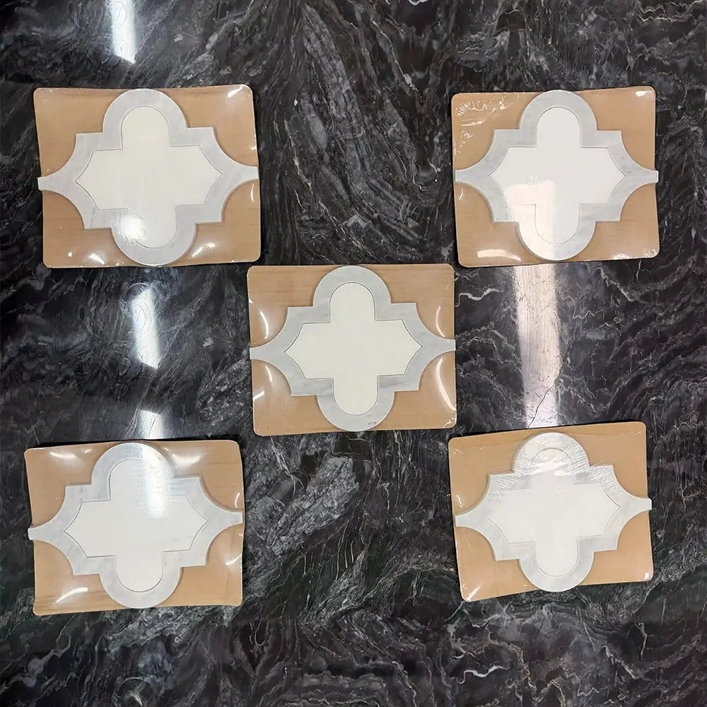 A flat-lay view of five Ares marble mosaic tile samples packaged on protective backing, showcasing the individual quatrefoil natural stone units on a dark marble surface.