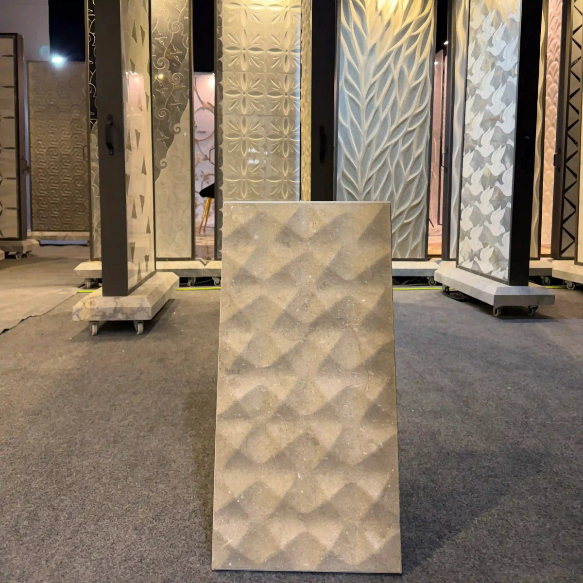 Echo natural stone 3D wall panel displayed as an individual vertical rectangular sample on a showroom floor, featuring a geometric faceted diamond or pyramidal textured pattern in a warm beige stone finish.