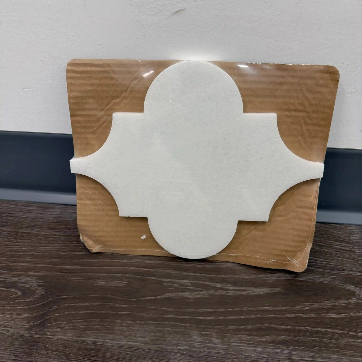 Helios natural marble mosaic tile presented as an individual sample piece on a dark wood-grain surface, featuring a classic, rounded quatrefoil or lantern silhouette in a smooth, uniform white marble.
