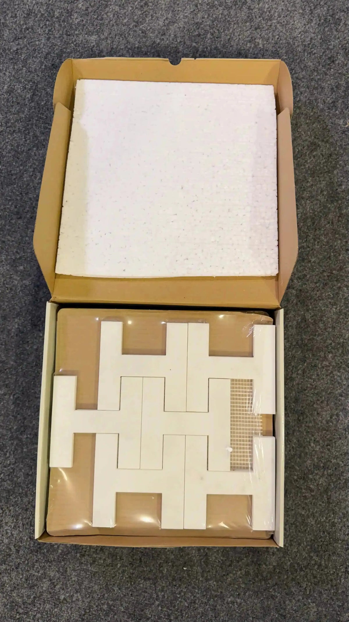 Icarus natural stone mosaic tile packaged securely inside an open cardboard box with protective foam inserts, showcasing interlocking H-shaped white marble pieces mounted on a mesh backing.