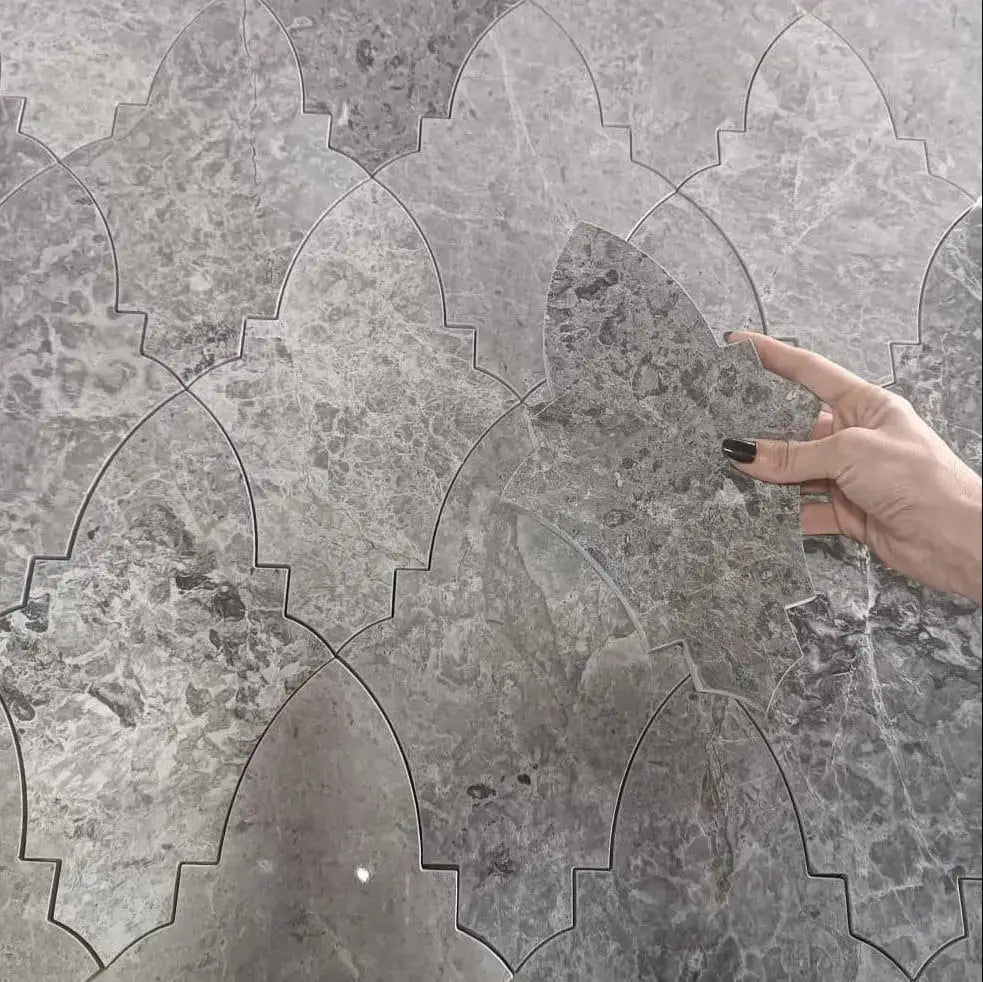 Khepri marble mosaic tile installation showing an intricate interlocking pattern of grey natural stone pieces, with a hand holding an individual tile unit against the tiled wall background.