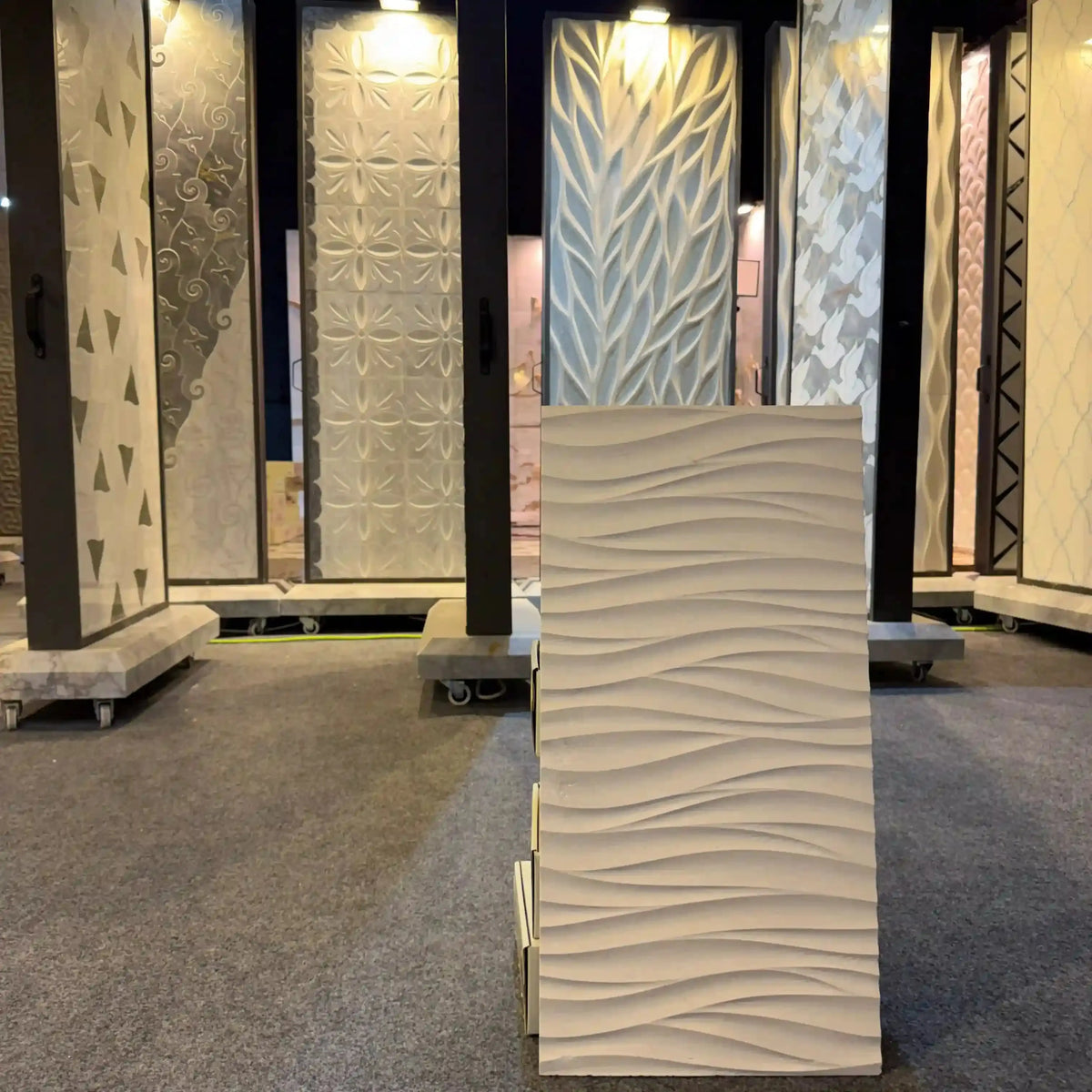 Medea natural stone 3D wall panel displayed as an individual vertical rectangular sample on a showroom floor, featuring a horizontal rolling wave-like sculpted texture in a neutral off-white or beige finish.